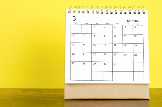 The March 2022 desk calendar with yellow background on the wooden table.