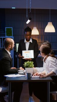 Overworked workaholic businesswoman with dark skin explaining management graphs using tablet overworking in company meeting office room late at night. Multi-ethnic teamwork solving company problem