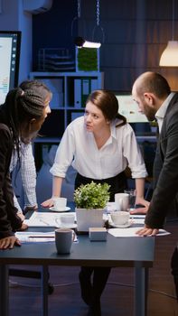 In corporate meeting room diverse multi ethnic group of businesspeople lean on conference table brainstorming project ideas late at night. Teamwork overworking at management presentation