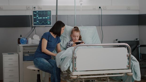 Mother sitting beside sick daughter while playing online video games streaming gamer app using smartphone. Little child with nasal tube resting in bed during medical examination in hospital ward