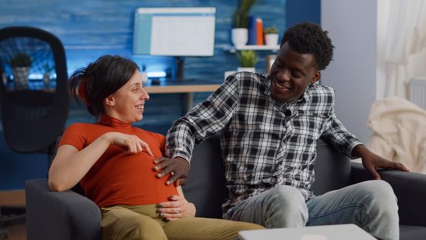 Married interracial couple expecting child sitting at home bonding on living room sofa. Caucasian mother laughing and relaxing while african american father of baby touching belly