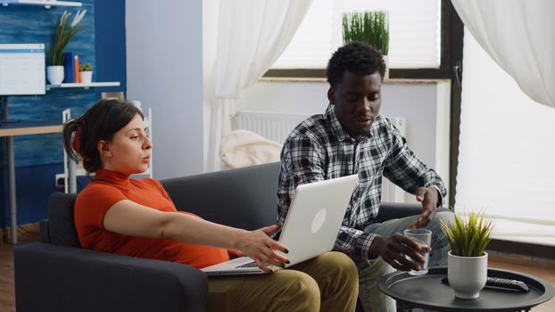 Interracial couple with pregnancy sitting together on couch while talking about child. Young pregnant woman using laptop and black man expecting baby and bonding in living room.