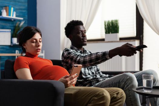 Young interracial couple expecting child at home. Pregnant caucasian woman looking at modern smartphone while african american man watching television with TV remote control in hand