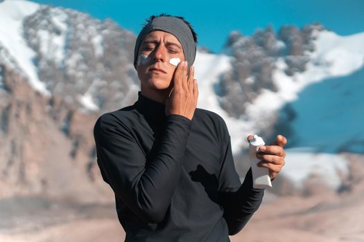 A young man applies sunscreen to his face in the highlands, in the background the peaks of the mountains are covered with snow. The traveler takes care of his skin while climbing.