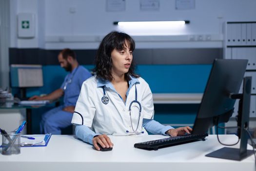Medical specialist using computer and keyboard at night, working late. Physician looking at monitor display for healthcare system and assistance after hours. Woman working as medic