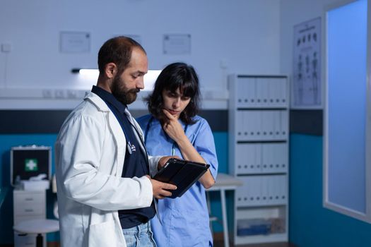 Doctor and nurse working late at night for healthcare using digital tablet. Medical assistant and medic doing teamwork at office for medicine. Woman and man looking at modern gadget