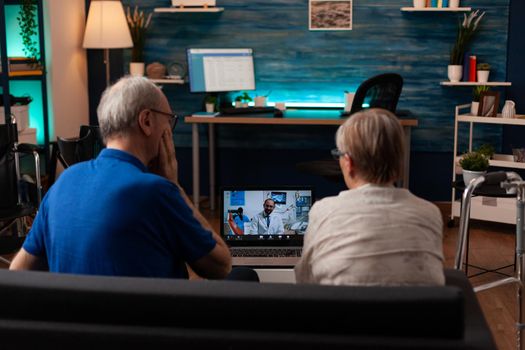 Senior couple talking on video call with dentist using laptop at home. Retired old people doing dentistry consultation, telemedicine with specialist while sitting in living room.