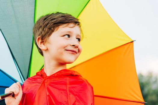 Portrait of a laughing school boy with rainbow umbrella behind. Smiling kid holds colourful umbrella on his shoulder. Cheerful child in a red raincoat holding multicolor umbrella.