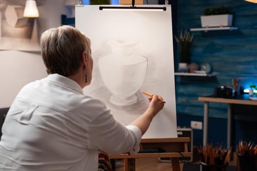 Close up of elderly woman drawing vase on canvas with pencil in workshop studio. Caucasian senior artist using object and artistic intruments for masterpiece artwork creation.