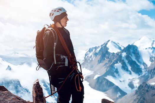 A young man traveler is engaged in mountaineering. In a helmet, with a rope, a harness, gloves, climbs to the top, against the backdrop of a stunning view with snow-capped mountains.