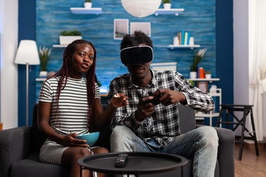 POV of black couple using technology for entertainment at home. African american man wearing vr glasses headset and playing with joystick while young woman watching video game on television