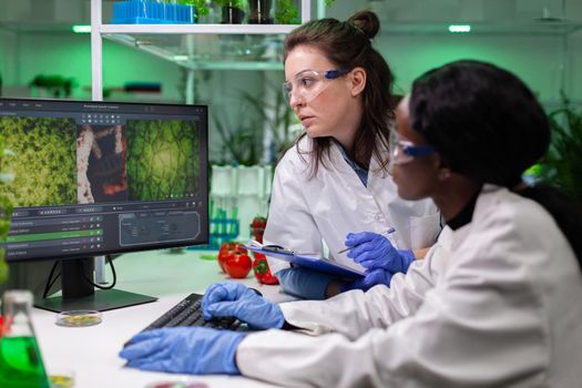 Multi-ethnic group of biologists analyzing plant based experiment on computer monitoring gmo test expertise. Diverse chemist team discovering genetic mutation in biochemistry hospital laboratory