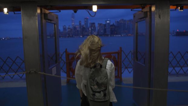 A female looks off into the distance, deep in thought while staring at the New York City skyline while riding the Staten Island Ferry. Back view of tourist on Staten Island Ferry approaching Manhattan in the evening.