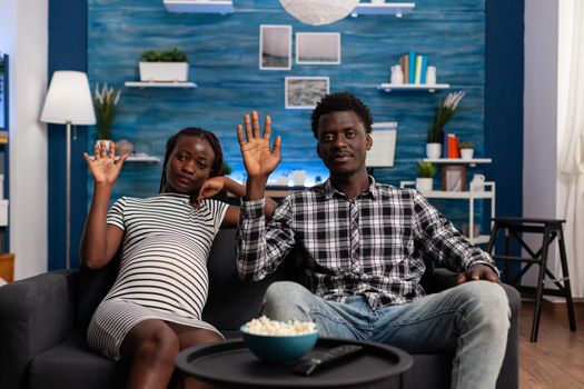 Pregnant black couple waving at video call camera and talking to friends sitting at home. People of african ethnicity expecting child while using online technology for remote communication