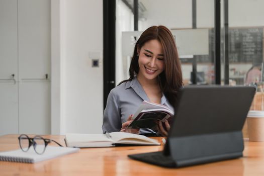 Smiling asian woman reading a book while studying or webinar online via tablet, video conference concept.
