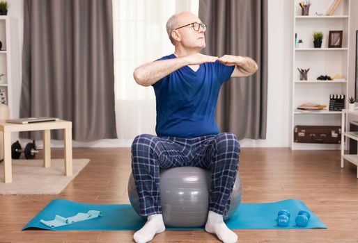 Active old man doing sport in his cozy apartment using swiss ball and yoga mat. Old person pensioner online internet exercise training at home sport activity with dumbbell, resistance band, swiss ball at elderly retirement age.