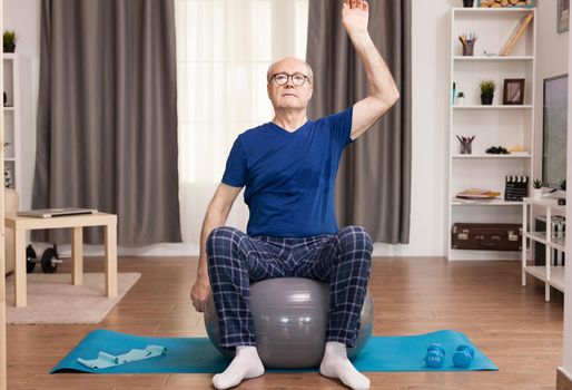 Elderly man training at home, doing arm exercises. Old person pensioner online internet exercise training at home sport activity with dumbbell, resistance band, swiss ball at elderly retirement age.