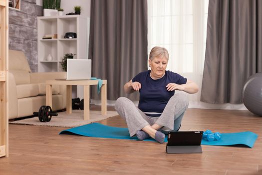 Retired woman warming up her shoulders before starting the fitness training. Old person pensioner online internet exercise training at home sport activity with dumbbell, resistance band