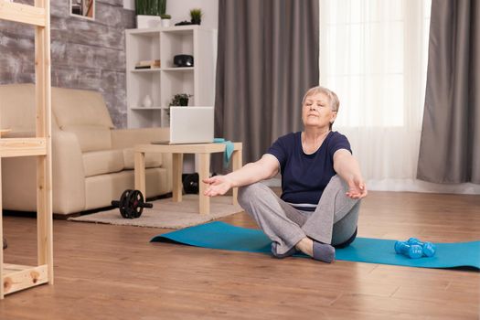 Elderly woman meditating in her cozy apartment. Old person pensioner online internet exercise training at home sport activity with dumbbell, resistance band, swiss ball at elderly retirement age