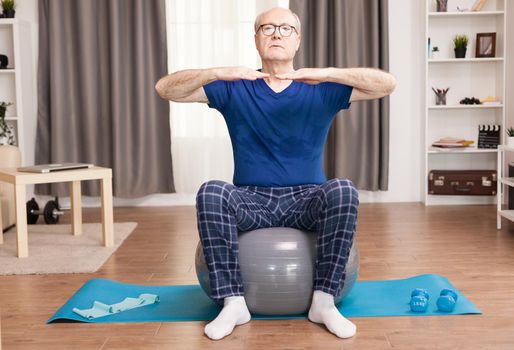 Senior man training on stability ball in living room. Old person pensioner online internet exercise training at home sport activity with dumbbell, resistance band, swiss ball at elderly retirement age.