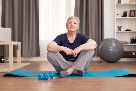 Portrait of elderly woman sitting on yoga mat. Old person pensioner online internet exercise training at home sport activity with dumbbell, resistance band, swiss ball at elderly retirement age