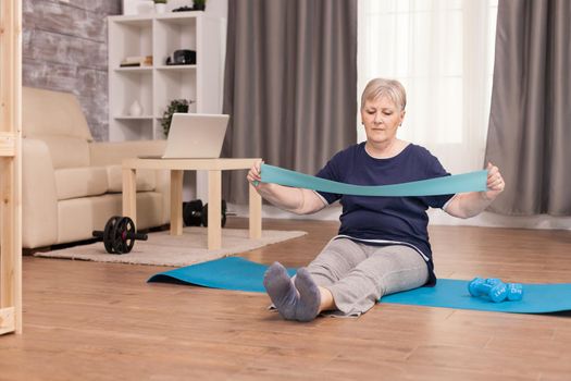 Active grandma using resistance band sitting on yoga mat at home. Old woman lifting training healthy lifestyle sport fitness workout at home with weights dumbbell activity.