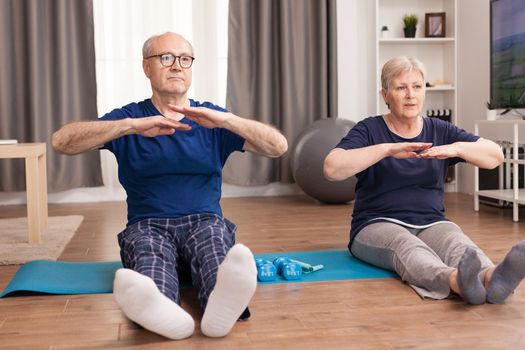 Senior couple stretching their bodies on yoga mat in living room. Old person healthy lifestyle exercise at home, workout and training, sport activity at home on yoga mat.