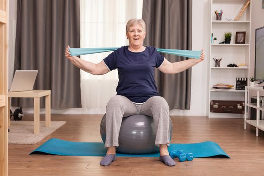 Aged woman enjoying exercise in her cozy apartment. Old person pensioner online internet exercise training at home sport activity with dumbbell, resistance band, swiss ball at elderly retirement age