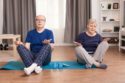 Senior couple relaxing sitting on yoga mat in the middle of the room. Old person healthy lifestyle exercise at home, workout and training, sport activity at home on yoga mat.