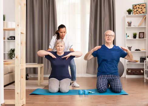Senior couple exercising with therapist on yoga mat at home. Home assistance, physiotherapy, healthy lifestyle for old person, training and healthy lifestyle