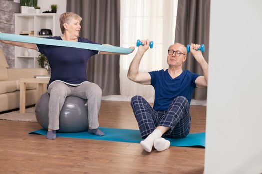 Elderly couple workout at home with elastic band and dumbbells. Old person healthy lifestyle exercise at home, workout and training, sport activity at home on yoga mat.
