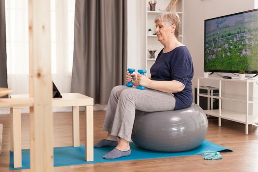 Senior woman training with weights watching online fitness lesson. Old person pensioner online internet exercise training at home sport activity with dumbbell, resistance band, swiss ball at elderly retirement age