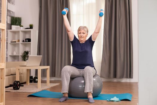Senior woman doing warm up exercise for recovery. Old person pensioner online internet exercise training at home sport activity with dumbbell, resistance band, swiss ball at elderly retirement age