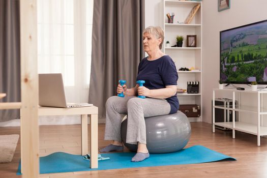 Old housewife watching online fitness lesson at home. Old person pensioner online internet exercise training at home sport activity with dumbbell, resistance band, swiss ball at elderly retirement age