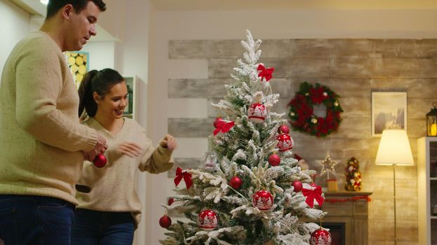 Couple with matching clothes decorate their christmas tree for winter holidays. Decorating beautiful xmas tree with glass ball decorations. Wife and husband in matching clothes helping ornate home with garland lights