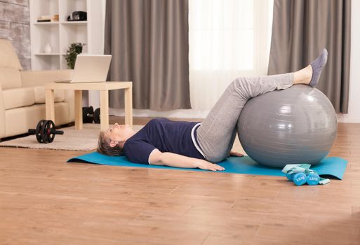 Old woman sitting on the yoga mat using a swiss ball to relax her legs. Old person pensioner online internet exercise training at home sport activity with dumbbell, resistance band, swiss ball at elderly retirement age