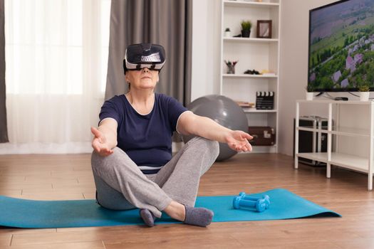 Elderly woman meditating with VR headset in the middle of the room. Old person pensioner online internet exercise training at home sport activity with dumbbell, resistance band, swiss ball and VR headset at elderly retirement age