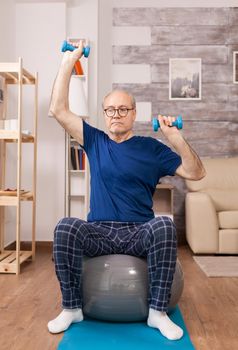 Elderly man doing sport in pajamas using dumbbells. Old person pensioner online internet exercise training at home sport activity with dumbbell, resistance band, swiss ball at elderly retirement age.