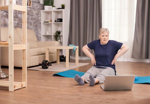 Senior woman warning up her shoulders looking on laptop. Old person pensioner online internet exercise training at home sport activity with dumbbell, resistance band, swiss ball at elderly retirement age