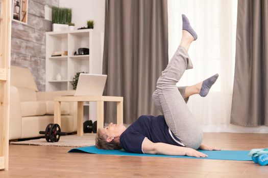 Old woman exercising in her apartment sitting on yoga mat. Old person pensioner online internet exercise training at home sport activity with dumbbell, resistance band, swiss ball at elderly retirement age