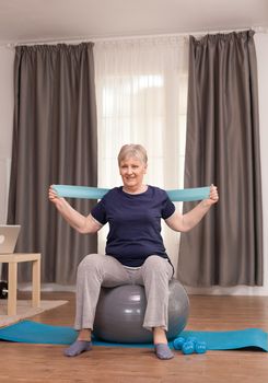 Modern senior woman training with resistance band sitting on balance ball. Old person pensioner online internet exercise training at home sport activity with dumbbell, swiss ball at elderly retirement age