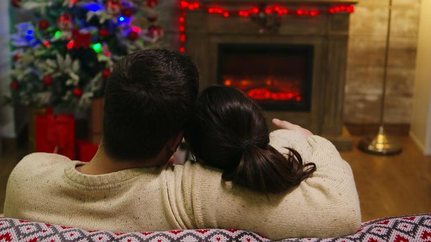 Back view of beautiful young couple sitting on couch looking at each other. Xmas spirit for caucasian wife and husband celebrating positive happy new year eve sitting whispering merry christmas