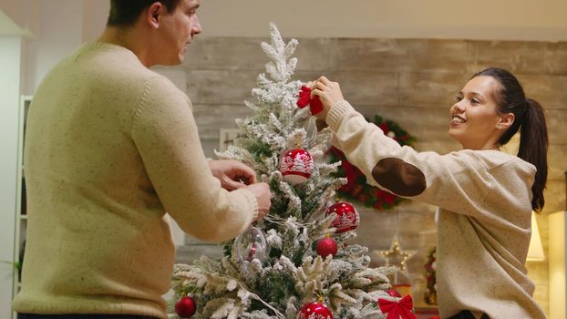 Caucasian man and woman happy to decorate their christmas tree. Winter holidays. Decorating beautiful xmas tree with glass ball decorations. Wife and husband in matching clothes helping ornate home with garland lights