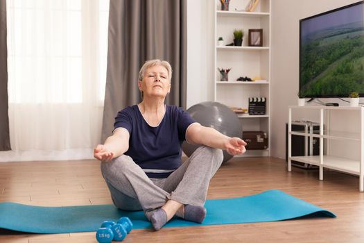 Old woman meditating on yoga mat at homeOld person pensioner online internet exercise training at home sport activity with dumbbell, resistance band, swiss ball at elderly retirement age