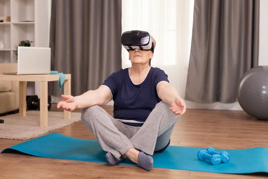 Grandmother wearing VR headset sitting on yoga mat. Old person pensioner online internet exercise training at home sport activity with dumbbell, resistance band, swiss ball and VR headset at elderly retirement age