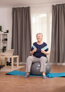 Healthy senior woman using resistance band for vitality. Old person pensioner online internet exercise training at home sport activity with dumbbell, swiss ball at elderly retirement age