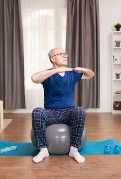 Retired man with healthy lifestyle doing arms exercises. Old person pensioner online internet exercise training at home sport activity with dumbbell, resistance band, swiss ball at elderly retirement age.