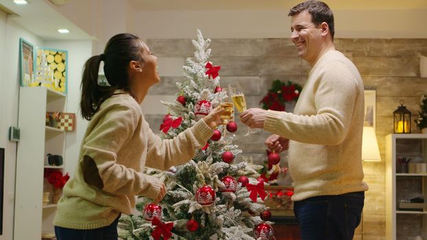 Caucasian man and woman happy to decorate their christmas tree. Winter holidays. Decorating beautiful xmas tree with glass ball decorations. Wife and husband in matching clothes helping ornate home with garland lights