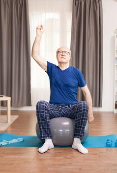 Old man training his arms sitting on stability ball in living room. Old person pensioner online internet exercise training at home sport activity with dumbbell, resistance band, swiss ball at elderly retirement age.