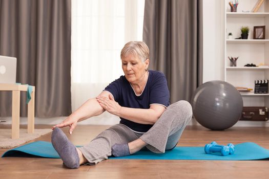 Active old woman exercising on the yoga mat in her comfortable apartment. Old person pensioner online internet exercise training at home sport activity with dumbbell, resistance band, swiss ball at elderly retirement age